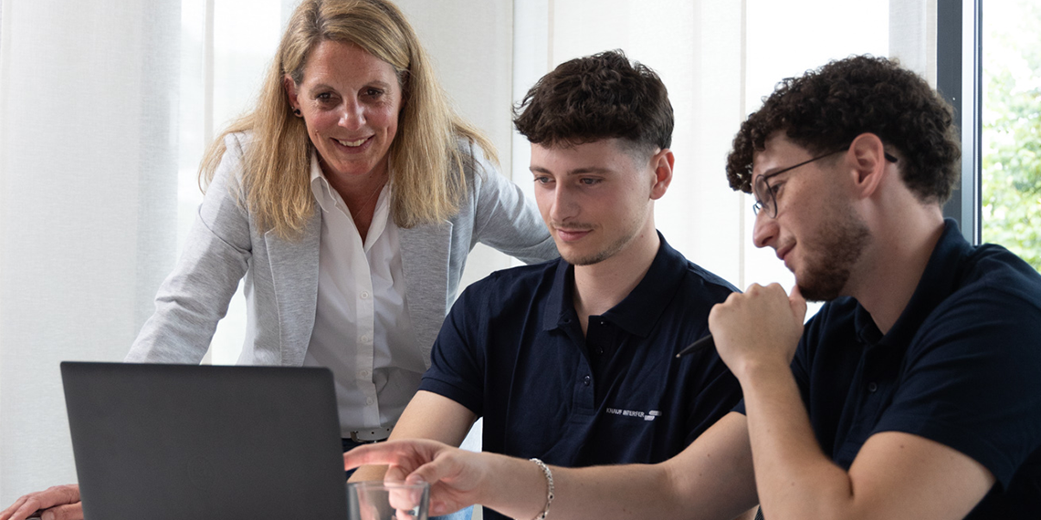 2 junge Auszubildende arbeiten am Laptop, einer von ihnen deutet auf den Bildschirm. Ihre Ausbilderin lächelt, während sie sie beobachtet.